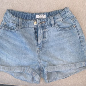 Ljusblå jeansshorts från Lindex - Snygga ljusblå jeansshorts från Lindex med klassisk femficksdesign och uppvikta benslut. Shortsen har normal passform, bälteshällor och dragkedja med knapp framtill. Perfekta för varma dagar och enkel att matcha med allt.