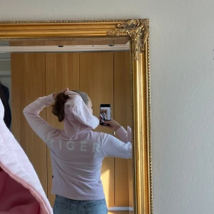 Ljusrosa hoodie från Tiger of Sweden - Ljusrosa hoodie från Tiger of Sweden Jeans med stor huva, snörning och magficka. Trycket 'TIGER' i vitt på ryggen ger en clean vibe. Mjuk bomullskänsla och ribbade muddar. Perfekt för chill dagar eller när du vill ha en enkel men snygg look.