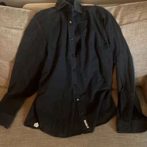 Svart skjorta från La Chemise - Stilren svart skjorta från La Chemise i slim fit-modell. Skjortan har klassisk krage, långa ärmar och knäppning framtill. Perfekt för dig som gillar en clean och enkel look. Materialet känns mjukt och lätt, troligen bomull.