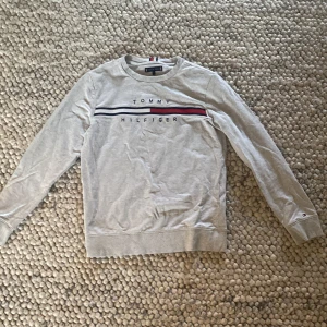 Grå Tommy Hilfiger sweatshirt(152 cm) - Snygg grå sweatshirt från Tommy Hilfiger med broderad logga och klassiska ränder i rött, vitt och marinblått över bröstet. Tröjan har rund hals, långa ärmar och är gjord i mjuk bomull. Perfekt för en chill och stilren look.