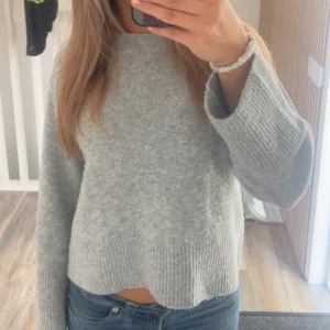 Grå stickad tröja med vida ärmar - Mysig grå stickad tröja med breda, långa ärmar och ribbade detaljer vid ärmslut och nederkant. Tröjan har en något croppad passform och är perfekt för dig som gillar en avslappnad och trendig stil.