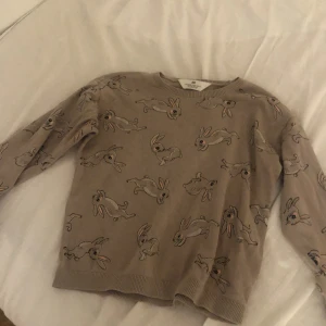 Beige tröja med kaninmönster H&M - Supersöt beige tröja från H&M med gulligt kaninmönster över hela plagget. Tröjan har rund halsringning, långa ärmar och ribbade muddar vid ärmslut och nederkant. Perfekt för dig som gillar djur och vill ha en mysig och lekfull stil.