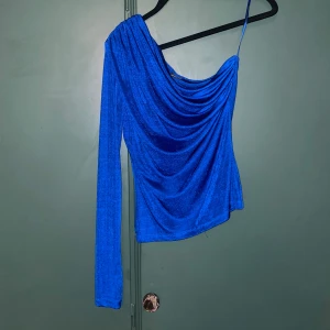 Blå oneshoulder topp Gina Tricot - Säljer en glittrig blå oneshoulder topp från Gina Tricot. Toppen har en lång ärm på ena sidan och med snygg drapering framtill. Använd 1-2 ggr