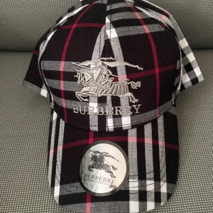 Burberry keps - Snygg keps från Burberry med klassiskt rutig mönster i svart, vitt, grått och röda detaljer. Broderad Burberry-logga och riddare framtill samt bak. Justerbar rem baktill. Perfekt accessoar för att lyfta din stil. Paket storlek=den får plats i en sko låda