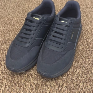 Svarta sneakers från Axel Arigato - Stilrena svarta sneakers från Axel Arigato med diskreta gulddetaljer på sidan och plösen. Skorna har en mix av mocka och textil, rund tå och platt sula. Perfekta för dig som gillar minimalistisk och modern design.
