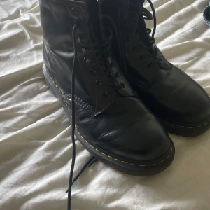 Svarta Dr. Martens boots - Klassiska svarta Dr. Martens boots i skinn med rund tå och snörning. Snygga gula sömmar runt sulan och dragflik baktill med logga. Perfekta för dig som vill ha en tidlös och edgy stil. Platt sula och robust känsla.