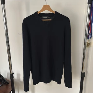 Svart stickad tröja från Dressmann - En klassisk svart stickad tröja från Dressmann med v-ringad hals och långa ärmar. Perfekt för lager-på-lager och enkel att matcha med jeans eller chinos. Tröjan har en normal passform och är tillverkad i mjukt material som känns skönt mot huden.