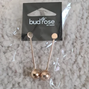 Guldiga statementörhängen Bud to Rose - Snygga och trendiga örhängen från Bud to Rose. De är guldiga med minimalistisk design, raka stavar med rund platta upptill och en större kula längst ner. Perfekta för dig som gillar stilrena smycken med en modern twist.