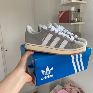 Grå Adidas Campus sneakers i mocka - Snygga Adidas Campus sneakers i grå mocka med klassiska vita ränder på sidorna och vit sula med beige kant. Skorna har rund tå, platt sula och breda vita snören. Perfekta för en clean och stilren look.