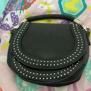 Gullig hanväska från carin Wester  - Snygg svart handväska från carin Wester med rundad form, dekorerad med silvriga nitar på framsidan och ett flätat handtag. Väskan är gjord i skinnimitation och har ett stilrent, trendigt uttryck. Perfekt för dig som vill ha en edgy accessoar till din outfit.