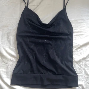 Linnetopp med öppen rygg - Säljer en svart linnetopp med tunna axelband och snygg draperad urringning. Toppen har glittriga detaljer i tyget och en djup öppen rygg med små strasspännen på banden. Perfekt för dig som vill sticka ut lite extra.