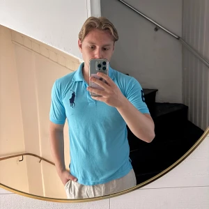 Ljusblå pikétröja från Polo Ralph Lauren - Säljer en ljusblå pikétröja från Polo Ralph Lauren i 100% bomull. Tröjan har klassisk krage, korta ärmar och stor mörkblå logga broderad på bröstet samt siffra på ärmen. Perfekt för en clean och sportig look. Sitter mer som S