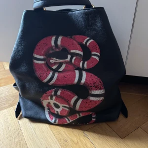 Gucci King Snake  ryggsäck med röd orm - Säljer en svart ryggsäck från Gucci i äkta skinn med en stor röd och vit orm på framsidan. Väskan har justerbara remmar, guldfärgade metalldetaljer och klassisk Gucci-tag inuti. Perfekt för dig som vill sticka ut med en unik och lyxig accessoar. Gucci by Alessandro Michele ”snake” collection. 