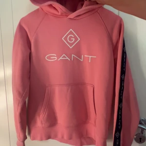 Rosa hoodie från GANT med logga - Säljer en rosa hoodie från GANT med vit logga framtill och svarta logoband längs ärmarna och huva. Tröjan har känguruficka och är långärmad med normal passform.