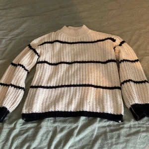 Vit och svart randig stickad tröja - Säljer en chunky stickad tröja i vitt med svarta ränder. Tröjan har hög krage och långa ärmar med breda muddar. Perfekt för kyliga dagar när du vill vara både bekväm och snygg. Passar dig som gillar en avslappnad och trendig stil.