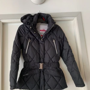 Svart quiltad dunjacka Röhnisch - Snygg svart dunjacka från Röhnisch med quiltat mönster, avtagbar huva och markerad midja med bälte. Jackan har dragkedjor på fickorna och en cool metallspänne framtill. Perfekt för kalla dagar när du vill vara både varm och stilren.