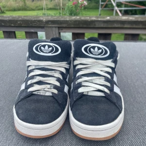 Adidas Campus svarta sneakers mocka - Svarta Adidas Campus sneakers i mocka med klassiska vita ränder på sidorna och vit Adidas-logga på plös och häl. Skorna har vit sula med gummidetalj och snörning framtill. Perfekt för dig som gillar retrostil och streetwear. Kommer att tvättas inan de skickas 