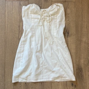Vit kort klänning från Bershka - Supersöt vit kort klänning från Bershka med snörning framtill och axelbandslös design. Klänningen har en figurnära passform och är tillverkad i ett lätt bomullstyg, perfekt för varma dagar. Smockad rygg för extra komfort och snygg siluett.