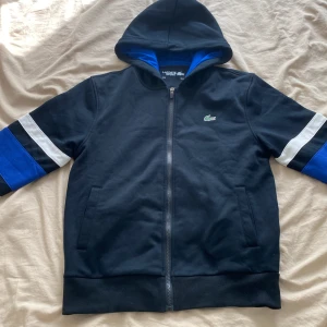 Lacoste Sport huvtröja - Svart hoodiejacka från Lacoste Sport med blå och vita detaljer på ärmarna. Jackan har dragkedja framtill, två fickor och en broderad grön krokodil på bröstet. Insidan av huvan är blå för en extra cool kontrast. Perfekt för en sportig och avslappnad stil. Saknar snören