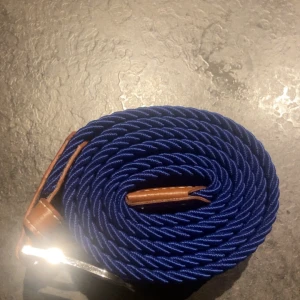 Blått flätat bälte med läderdetaljer - Snyggt blått flätat bälte med bruna läderdetaljer och silverfärgat spänne. Bältet har en elastisk känsla och passar perfekt till jeans eller chinos. Den flätade designen ger en cool och modern look.