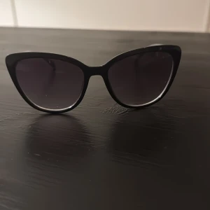 Svarta cat eye solglasögon - Snygga svarta solglasögon i klassisk cat eye-modell med mörka glas. Ramen är i blank plast och ger en cool och tidlös look. Perfekta för att lyfta vilken outfit som helst med lite extra attityd.