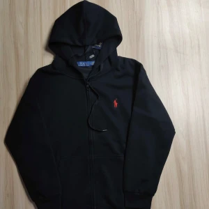 Svart hoodie från Polo Ralph Lauren - Svart hoodie från Polo Ralph Lauren med röd broderad logga på bröstet. Klassisk modell med dragkedja, huva med snörning och känguruficka. Tillverkad i mjuk bomull, perfekt för chill dagar eller när du vill ha en clean look. Den är även helt ny. 
