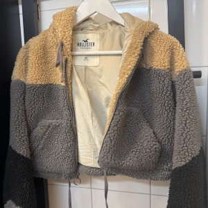 Teddyjacka från Hollister i beige och grå - Supermjuk teddyjacka från Hollister i färgblockad beige, grå och svart. Jackan har huva, dragkedja med ringdetalj och två fickor framtill. Perfekt för kyliga dagar och ger en chill vibe. Croppad modell som sitter snyggt till jeans eller mjukis.
