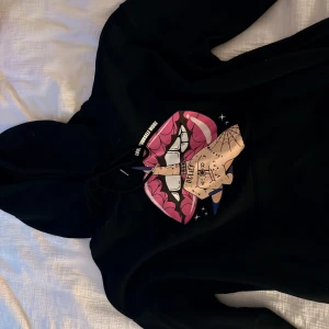 Svart hoodie med tryck och huva - Svart hoodie med stor färgglad print framtill av rosa läppar, tänder och en tatuerad hand med blå naglar. Texten 'LOVE YOURSELF MORE' och 'BELIEVE' syns i motivet. Tröjan har huva med snörning och är perfekt.