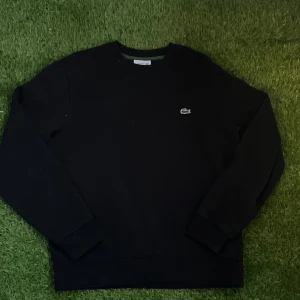 Svart sweatshirt från Lacoste - Klassisk svart sweatshirt från Lacoste i bomull med rund hals och broderad krokodil-logga på bröstet. Tröjan har långärmad passform och ribbade muddar vid ärmslut och nederkant. Perfekt för en clean och stilren look. Tvättar självklart innan ja skickar 