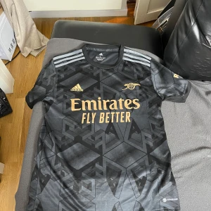 Arsenal bortatröja Adidas L svart/guld - Snygg Arsenal bortatröja från Adidas i storlek L. Tröjan är svart med geometriskt mönster och guldiga detaljer som klubbmärke, Adidas-logga och sponsortryck. Tillverkad i lätt och ventilerande Aeroready-material, perfekt för match eller träning.