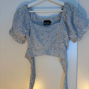 Blommig croppad topp från Pieces - Supersöt ljusblå croppad topp från Pieces med blommigt mönster i lila, rosa och gult. Puffiga korta ärmar med resår och volangdetalj, fyrkantig ringning och långa knytband i midjan. Perfekt för vår och sommar när du vill sticka ut lite extra.