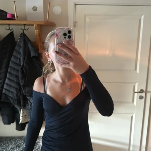 Svart omlottopp med offshoulder - Säljer en svart omlottopp med lång ärm och offshoulder-design. Toppen har smala axelband och en snygg v-ringning framtill. Materialet är stretchigt och sitter snyggt på kroppen. Perfekt för dig som gillar trendiga och stilrena plagg.