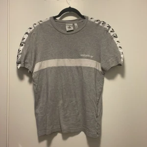 Grå Adidas t-shirt med vita detaljer - Snygg grå t-shirt från Adidas med vit rand över bröstet och vita axelband med svarta Adidas-loggor. Klassisk passform och korta ärmar, perfekt till streetwear eller träning. Mjukt material som känns skönt mot huden.