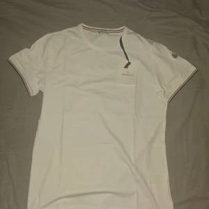 Vit slim fit t-shirt från Moncler - Vit slim fit t-shirt från Moncler med klassisk rund hals och korta ärmar. T-shirten har Moncler-logga på ärmen och snygga ränder i rött, vitt och blått på ärmslut och halsringning. Tillverkad i mjuk bomull för en clean och fräsch look. ------ Storlek är M/L (slim fit). 