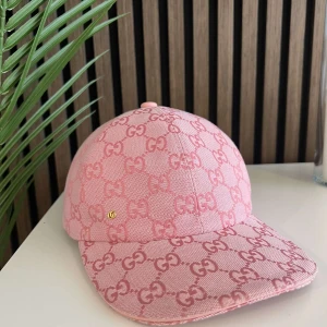 Rosa Gucci keps med GG-mönster - Snygg rosa keps från Gucci med ikoniskt GG-mönster över hela kepsen. Justerbar rem baktill i matchande rosa och liten guldfärgad GG-detalj framtill. Kepsen är tillverkad i ett slitstarkt bomullstyg och har en klassisk böjd skärm.