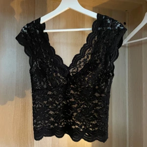 Svart spetsblus med v-ringning - Supersnygg svart blus i spets med djup v-ringning både fram och bak. Blusen är ärmlös med breda axelband och har vågiga kanter för en extra detalj🖤