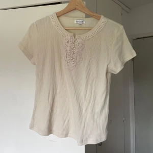 Beige ribbad topp med spetsdetalj - Säljer en beige ribbad topp med korta ärmar. Toppen har en rund halsringning med virkade spetsdetaljer och pärlor framtill som ger en söt vintagekänsla. Perfekt för dig som gillar romantiska detaljer och mjuka färger.