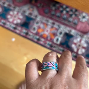 Färgglad bred ring med geometriskt mönster - Säljer en bred stål ring i silverfärgad stål med färgstarkt geometriskt mönster i blått, rosa och turkost. Ringen har coola trianglar och linjer som ger en unik och modern vibe. Perfekt statement piece för dig som gillar att sticka ut.