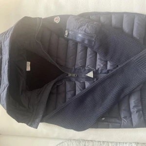 Moncler Cardigan - Snygg mörkblå pufferjacka från Moncler med stickade ärmar och detaljer. Jackan har dragkedja framtill, quiltad kropp och klassisk Moncler-logga på ärmen. Perfekt för dig som vill ha en stilren och exklusiv look under kyliga dagar.
