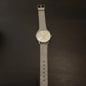 Silverfärgad Calvin Klein klocka - Stilren klocka från Calvin Klein med rund urtavla och mesh-armband i silverfärgat rostfritt stål. Minimalistisk design med diskret CK-logga på urtavlan och små markeringar för timmarna. Perfekt accessoar för en clean look. Batteri behöver bytas 