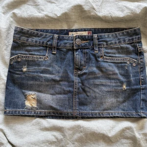 Jeanskjol från Fishbone, storlek S - Snygg blå jeanskjol från Fishbone i kort modell med slitna detaljer och klassiska fickor både fram och bak. Kjolen har bälteshällor, knapp och dragkedja framtill samt coola kontrastsömmar. Perfekt för dig som gillar en avslappnad och trendig stil.