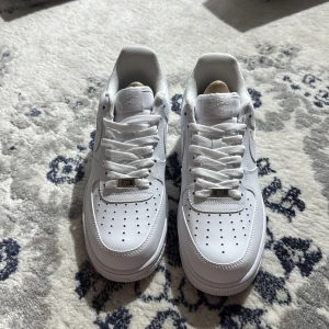 Nike Air Force 1 helvita sneakers - Klassiska Nike Air Force 1 i helvitt skinn med perforerad tå, rund tå och platt sula. Snygg och tidlös modell med snörning och diskret Nike-logga på sidan. Perfekta för dig som vill ha en clean och fräsch look till vardags.