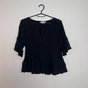Svart blus med volang från Vila - Svart blus från Vila med volangdetaljer och knytning vid halsen. Blusen har trekvartsärmar med lätt rynk och volangkant, samt en lös passform. Blusen har aldrig blivit använd för att den inte passade i storlek men den är väldigt fin. 