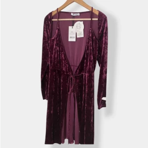 Mörklila morgonrock från NA-KD - Mörklila sammets morgonrock från NA-KD i modellen Velvet Overlap Dress. Morgonrocken har omlott-skärning, lång ärm och knytband i midjan. Materialet är mjuk sammet som ger en lyxig känsla och färgen är en djup lila nyans. Perfekt för dig som vill sticka ut.