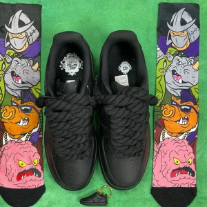 Nike Air Force 1 Low Triple Black - Nike Air Force 1 Low i helsvart med chunky snörning och klassisk siluett. Skorna är gjorda i skinn och har en platt sula samt rund tå. Perfekta för dig som gillar en clean och tidlös look. Säljes med coola färgglada strumpor med Turtles-motiv.