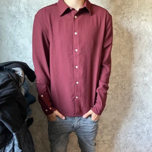 Vinröd skjorta från H&M Regular Fit - Vinröd skjorta från H&M i regular fit med klassisk krage och vita knappar framtill. Skjortan har lång ärm och en stilren, enkel design som passar till jeans eller chinos. Materialet känns lätt och har en diskret struktur för en snygg look. Pris ej hugget i sten vid snabb affär. hör av dig vid minsta fundering!