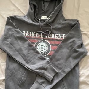 Grå hoodie från Saint Laurent - Snygg grå hoodie från Saint Laurent med tryck framtill i vitt och rött. Tröjan har huva med snörning och långa ärmar. Perfekt för dig som gillar streetstyle och vill ha något bekvämt men ändå stilrent.