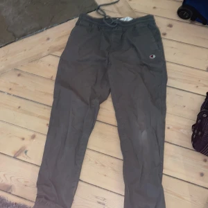 Gråa mjukisbyxor från Champion M - Gråa mjukisbyxor från Champion med elastisk midja och dragsko. Byxorna har en relaxed fit, resår vid benslut och Champion-logga på ena benet. Perfekta för chill eller träning. Tillverkade i mjukt material för extra komfort.