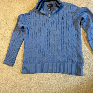 Blå stickad tröja Polo Ralph Lauren - Snygg blå kabelstickad tröja från Polo Ralph Lauren med halv dragkedja och ribbade muddar. Klassisk design med broderad logga på bröstet och hög krage med randig insida.  I princip o använd priset är inte hugget i sten 
