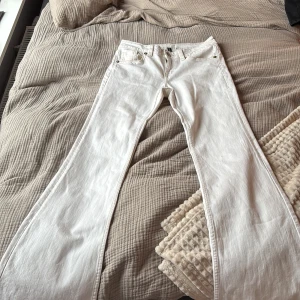 Vita bootcut jeans från H&M  - Snygga vita jeans från H&M med bootcut-modell och guldiga knappar. Byxorna har klassiska fem fickor och är tillverkade i ett mjukt bomullstyg. Perfekta för dig som vill ha en clean och fräsch look med lite extra edge.